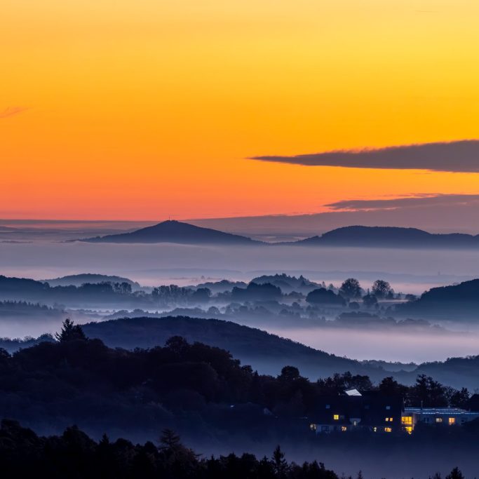 Sonnenuntergang über der Eifel, oranger Himmel. Nebelige Täler zwischen sanften Hügeln.